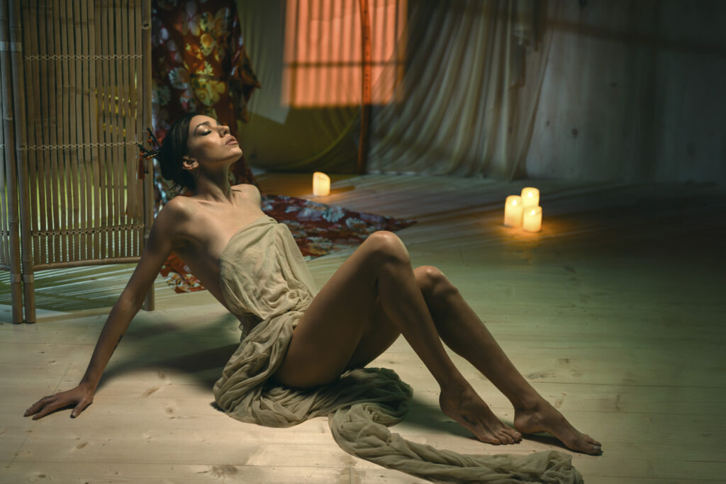 Modella coperta da un solo velo color carne in uno scenario Japponese orientale allestito per un set fotografico in stile cinematografico di stage photography realizzato da Francesco Francia ambassador nanlite e fotografo di moda. Luce in fotografia realizzata sul set con pochi elementi di scena, con una katana sullo sfondo , un mobile di bambù e delle candele con veli. luci cinematografiche in fotografia